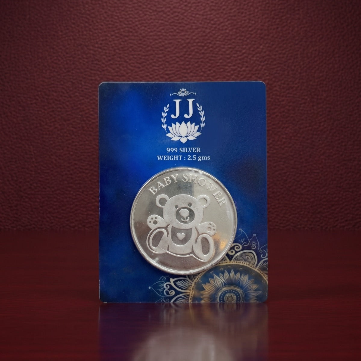 A silver baby shower coin featuring a teddy bear design, mounted on a blue card with 'JJ' branding, set against a dark red textured background.