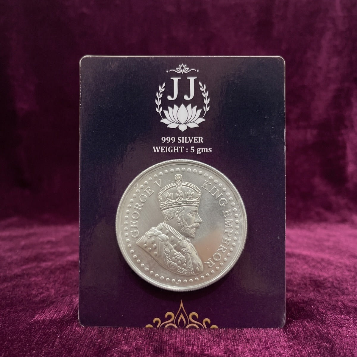 A silver coin mounted on a dark display card, photographed against a rich purple velvet fabric background. The card features the 'JJ' brand logo and text indicating it is 999 silver with a weight of 0.